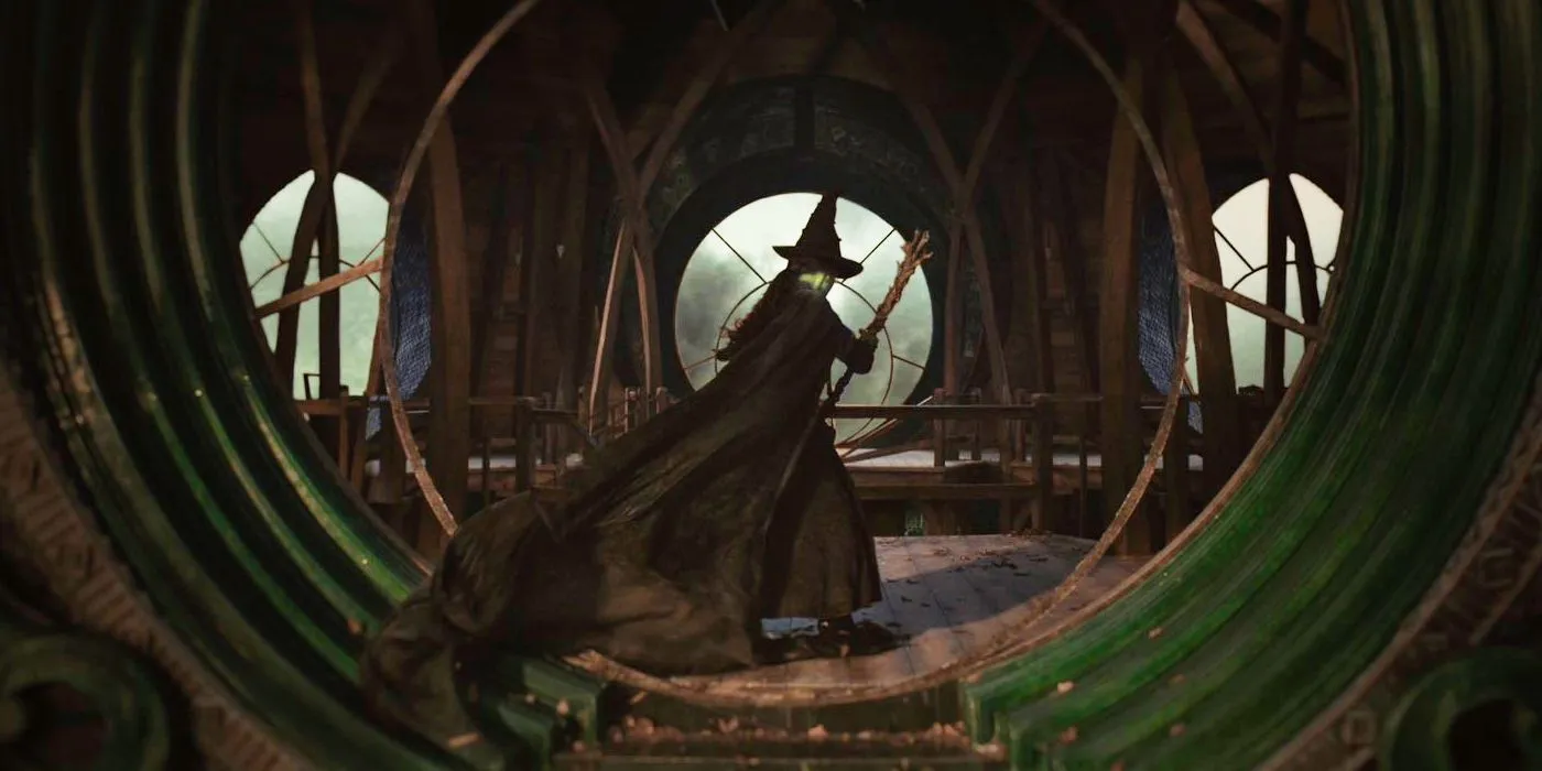 Elphaba با جارویش کنار پنجره شکسته در Wicked ایستاده است.